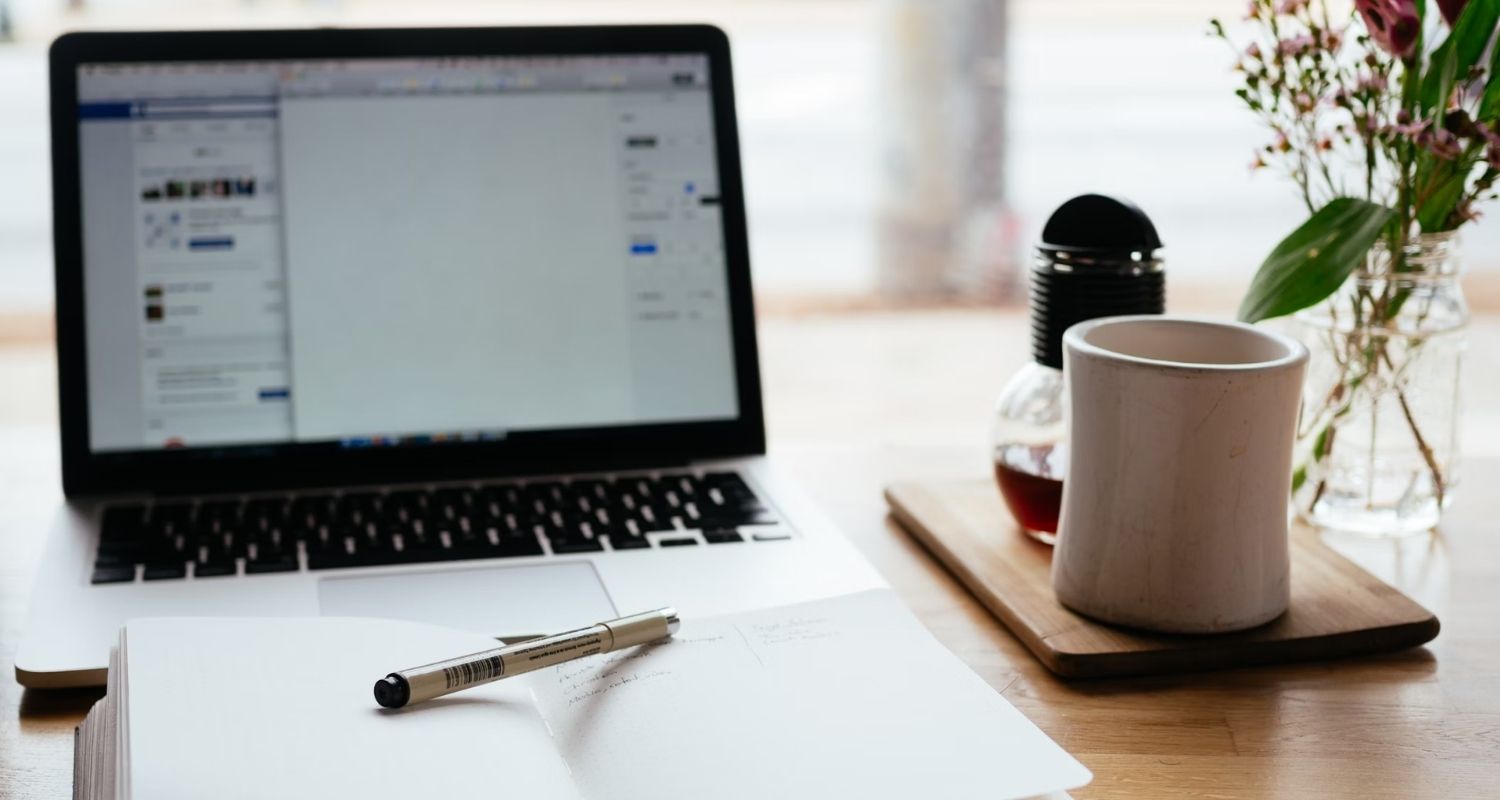 Open notebook with a pen beside a laptop displaying a document, next to a coffee mug and flowers on a wooden desk.