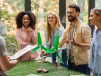 Team of young professionals discussing a miniature wind-turbine model in a sunlit office, showcasing corporate sustainability.