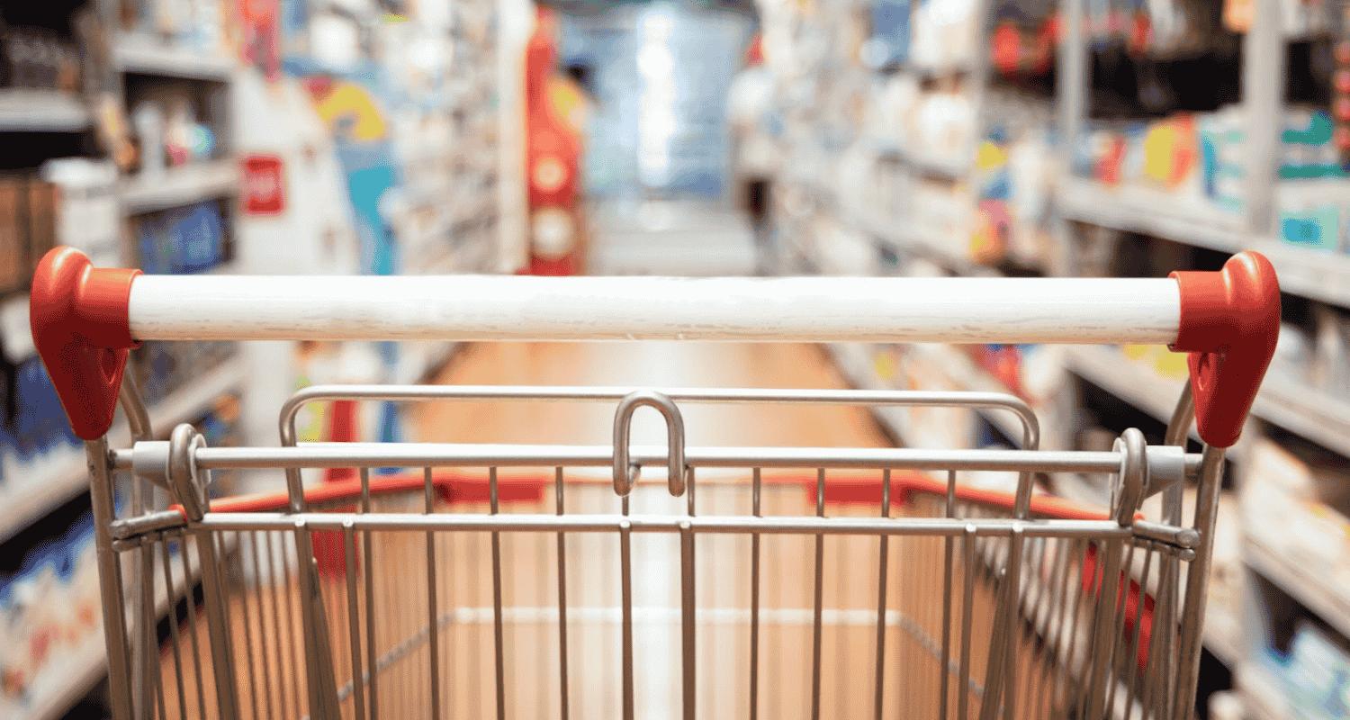 A shopping cart with a white handle and red grips is in focus, facing a brightly lit grocery aisle with blurred shelves stocked on both sides.