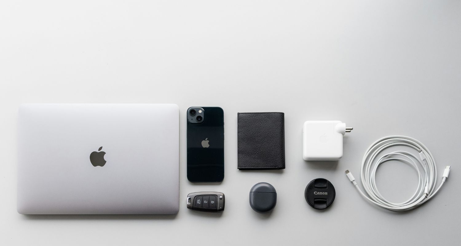 MacBook, iPhone, a wallet, charger, Car keys placed on a grey surface.