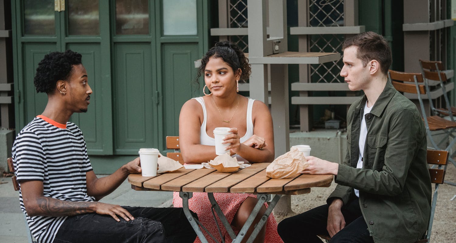 Three people sitting outdoors at a wooden table, enjoying coffee and engaged in a casual conversation.