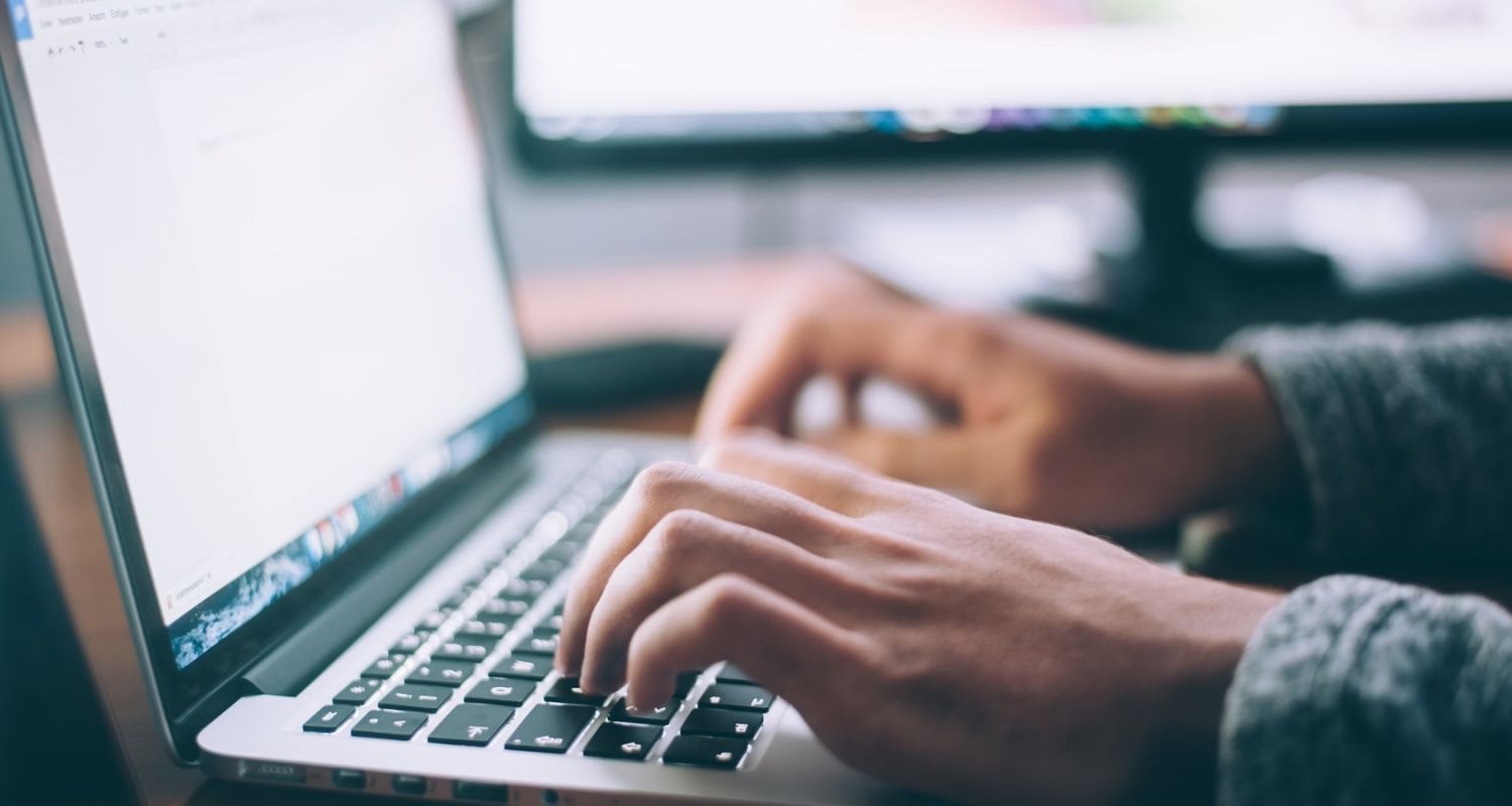 Close-up of hands typing on a laptop keyboard with a blurred screen and a monitor in the background.