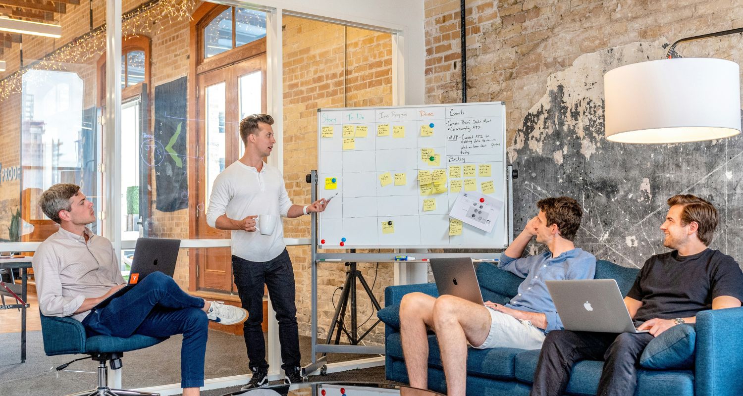 A team collaborating in the office, discussing ideas around a whiteboard filled with sticky notes, while working on laptops.