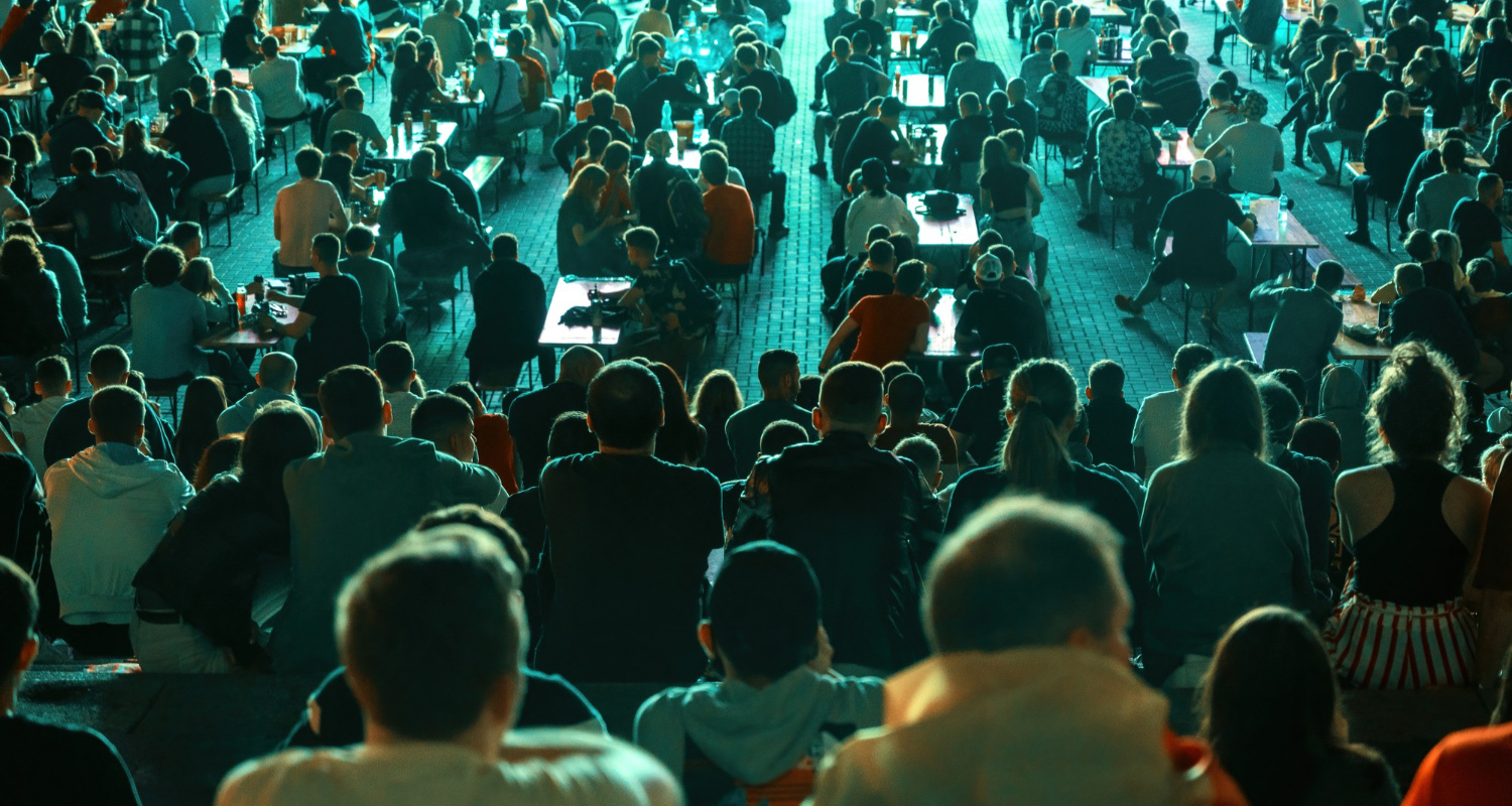 A large crowd at an outdoor event, seated at tables and benches, illuminated by blue-green lighting from behind.